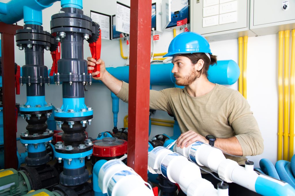 A technician in a blue hard hat is operating equipment and valves in a pool pump room with various pipes and machinery.