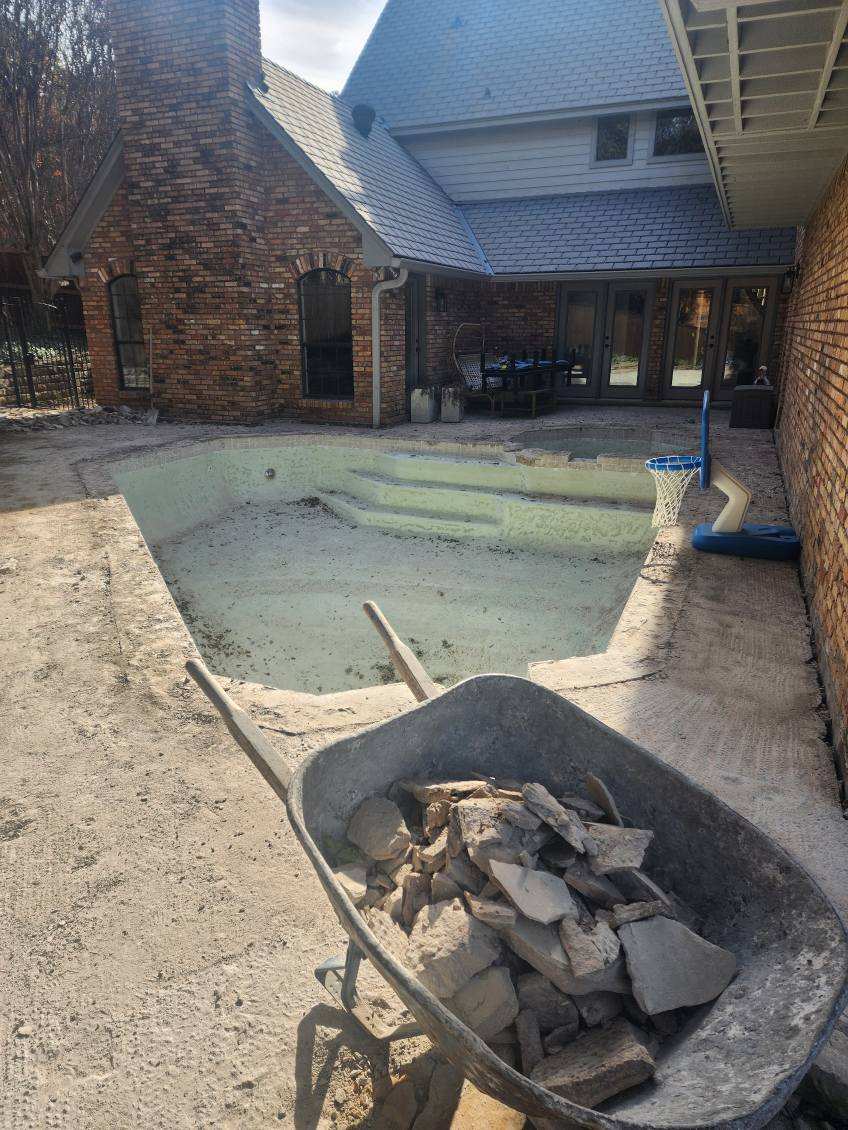 Construction site of a residential pool undergoing renovation, featuring a partially empty pool, a wheelbarrow filled with stones, and a brick house in the background.