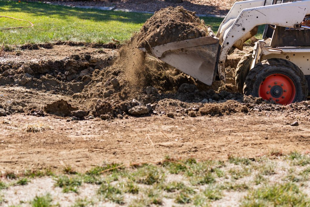 A backhoe excavator digging earth in a residential yard, creating a pit in the ground for pool removal or landscaping.
