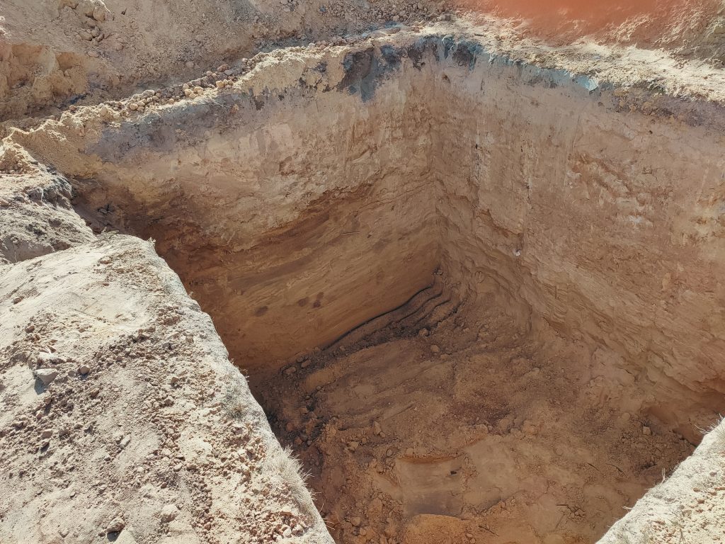 Excavated hole in the ground, showing layered soil and earth materials, likely for a pool removal project.