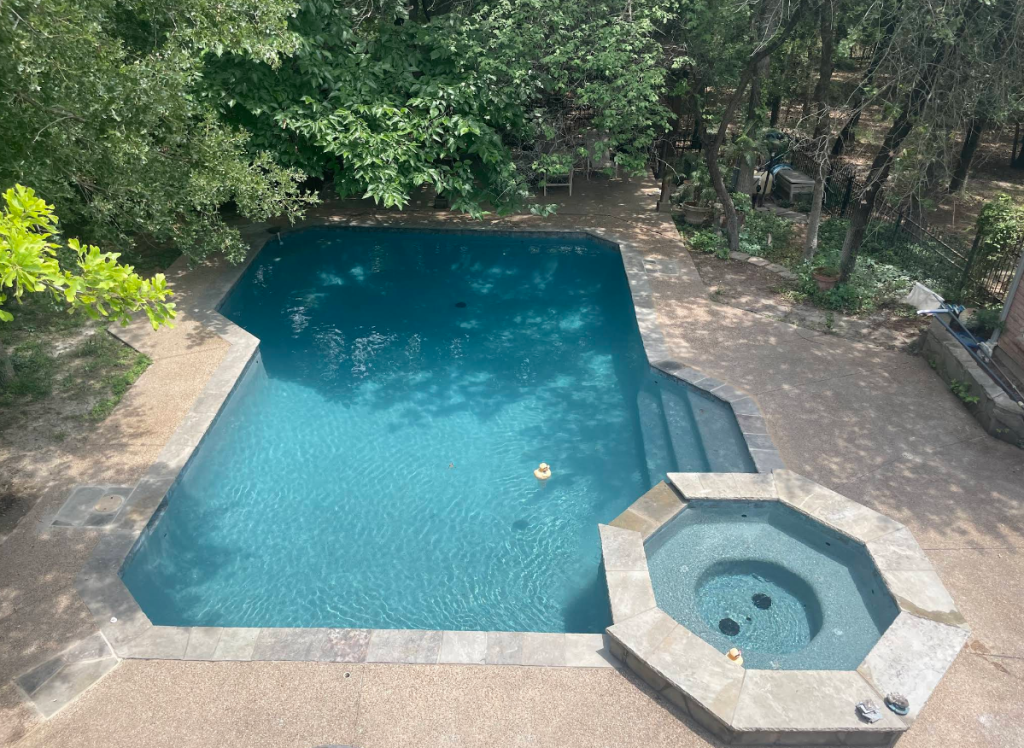 Aerial view of a rectangular swimming pool surrounded by trees, featuring a built-in spa with stone coping and a few decorative floating items in the water.