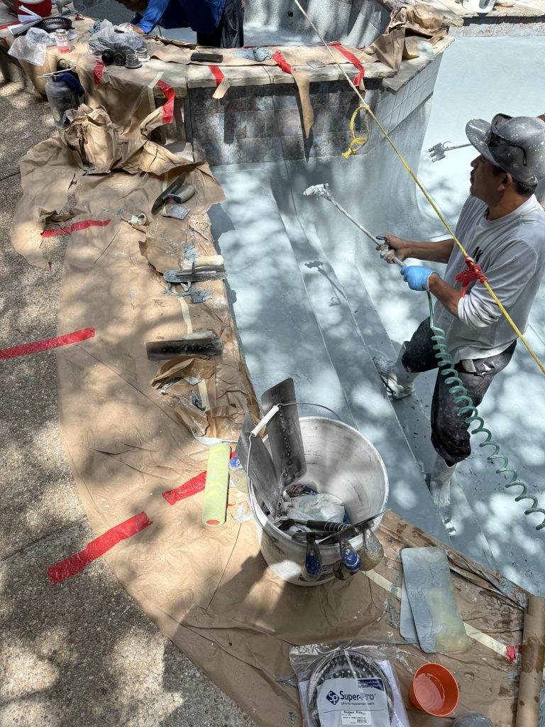Workers applying resurfacing materials inside a swimming pool under renovation, surrounded by tools and protective coverings.