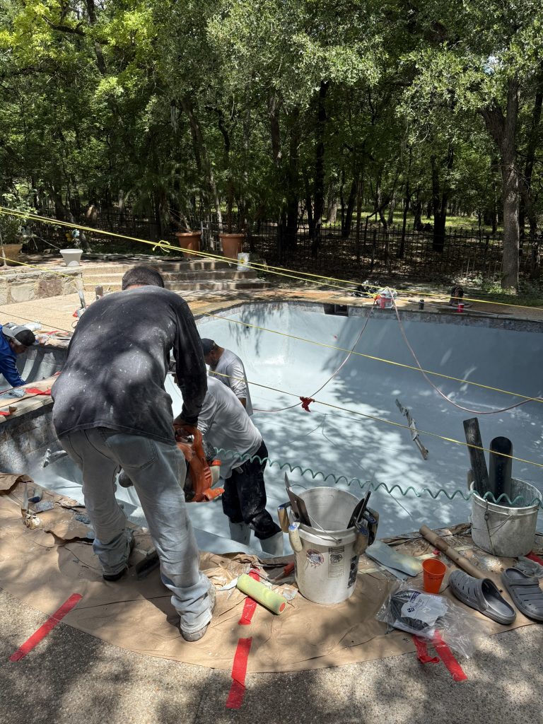 Workers performing renovations on a residential swimming pool, surrounded by trees, with construction tools and materials visible on the ground.