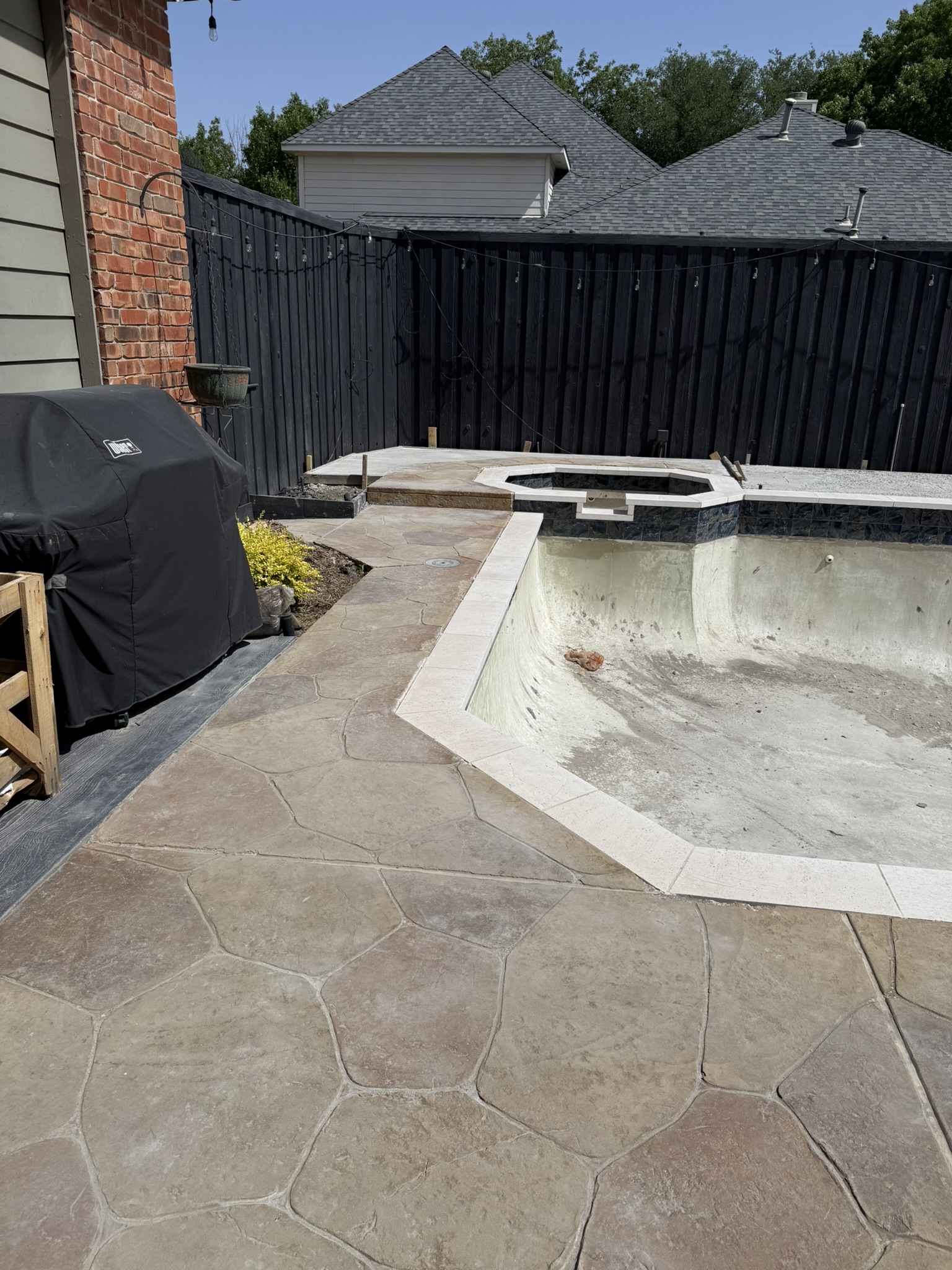 View of a partially completed pool remodel featuring a textured stone deck overlay, with an unfinished pool shell and rustic brick wall in the background.