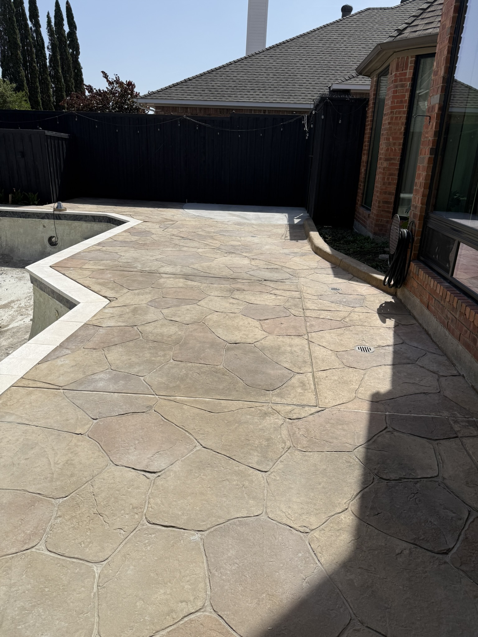 Outdoor patio area featuring a textured stone overlay with geometric patterns, alongside a swimming pool in a residential backyard.