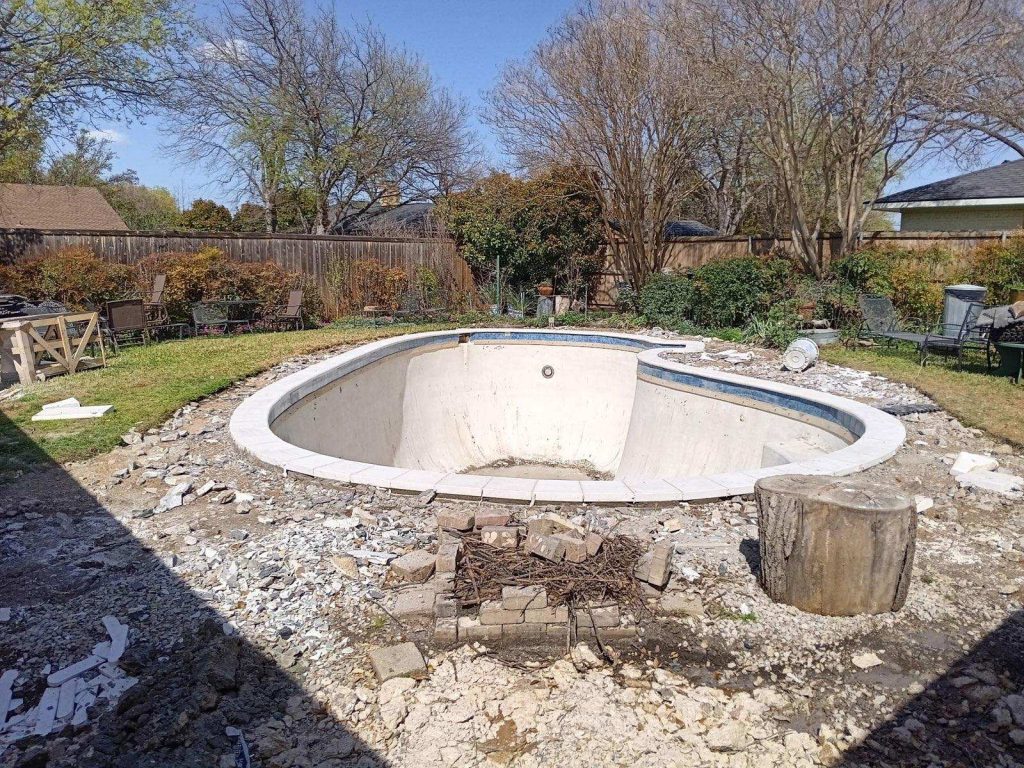 A partially demolished residential swimming pool in a backyard, showing exposed concrete and surrounding construction debris, with trees and a garden in the background.