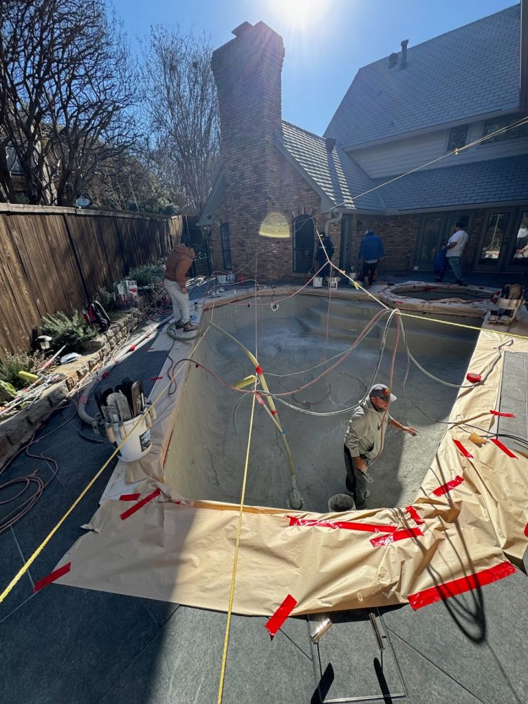 A construction worker applies material to a recently drained swimming pool in a backyard setting, with a house made of brick and a sloped roof in the background.