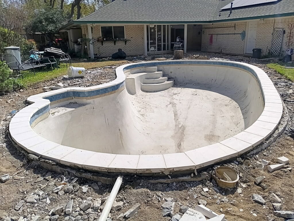 Empty swimming pool under renovation, featuring a tiled edge and steps, surrounded by construction debris and the backyard of a house.