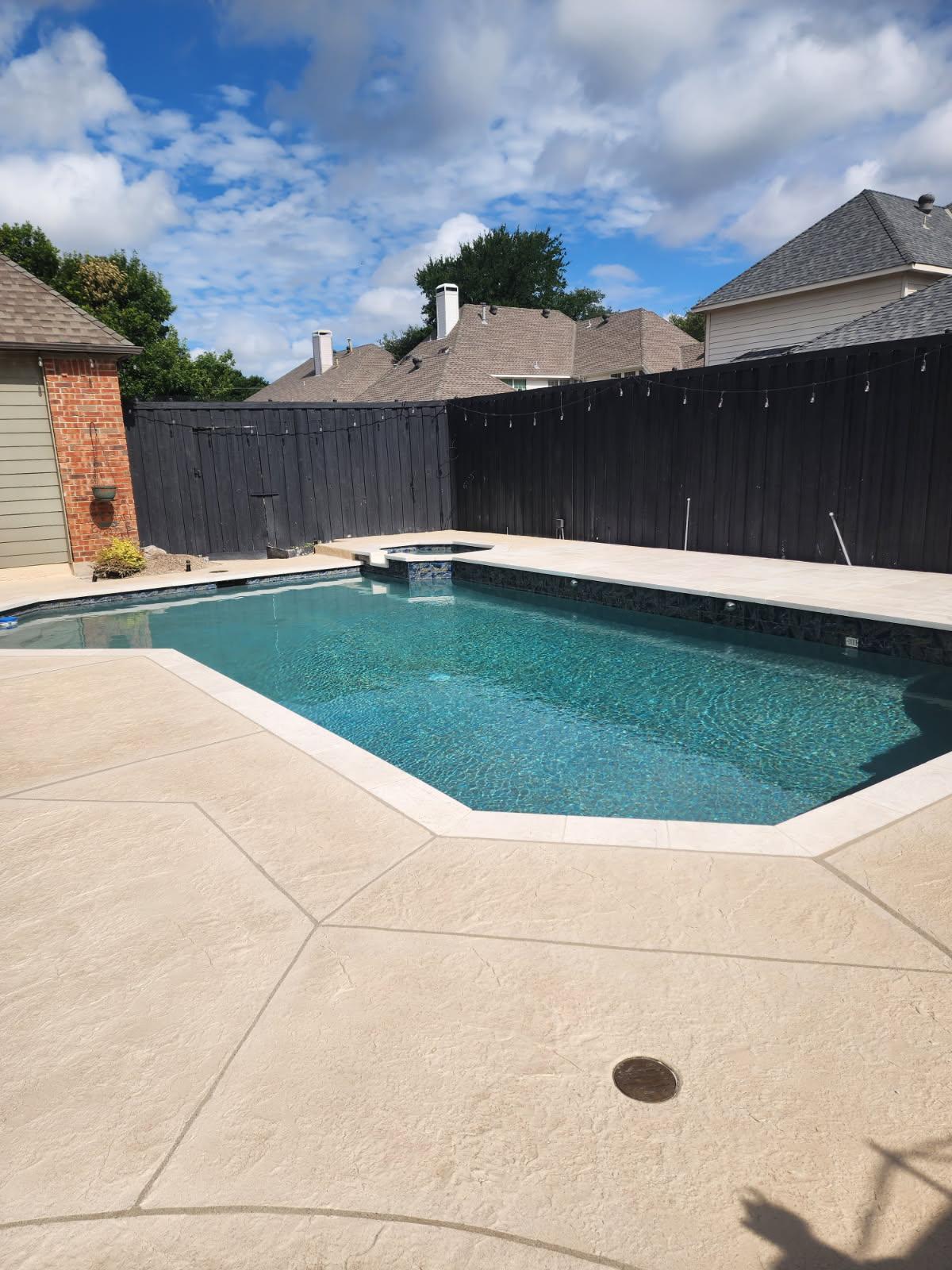 A renovated residential pool featuring a modern design with an octagonal shape, surrounded by a textured deck. The pool is clear and sparkling, with a backdrop of a wooden privacy fence and sunny blue skies.