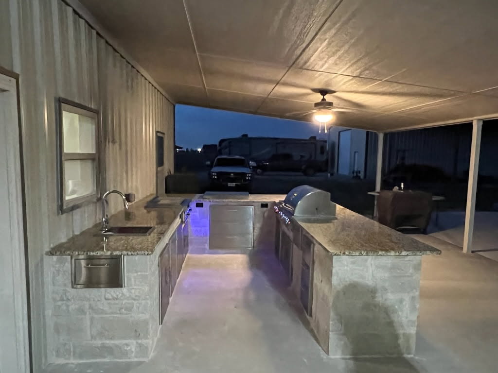 An outdoor kitchen setup featuring a granite countertop, stainless steel appliances, and an overhead ceiling fan, under a covered patio in a dimly lit setting.