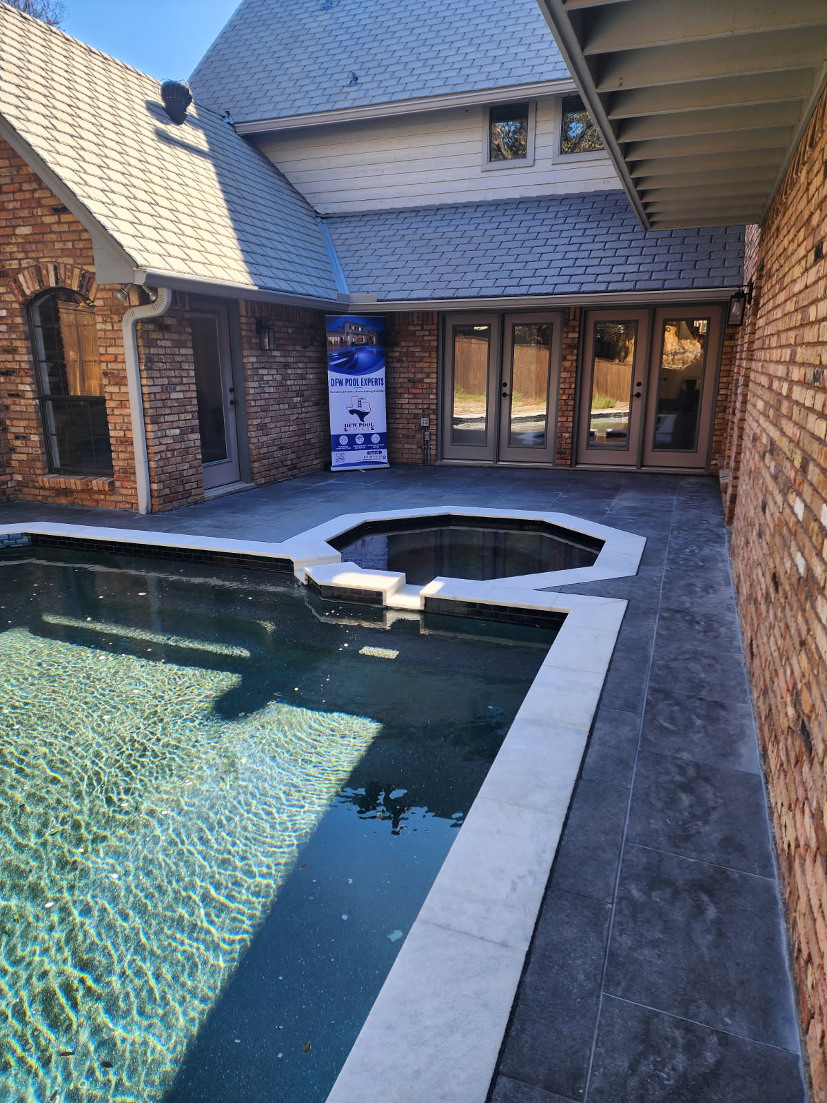 A recently remodeled residential pool area featuring clear water, modern deck tiling, and a spa section, set against the backdrop of a brick and shingle home.
