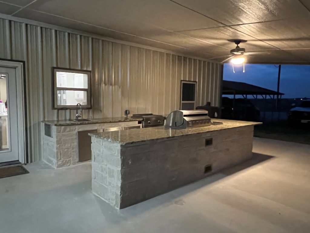 A modern outdoor kitchen featuring a stone countertop, built-in grill, and sink under a covered area with a fan, illuminated by soft lighting, creating an inviting ambiance.