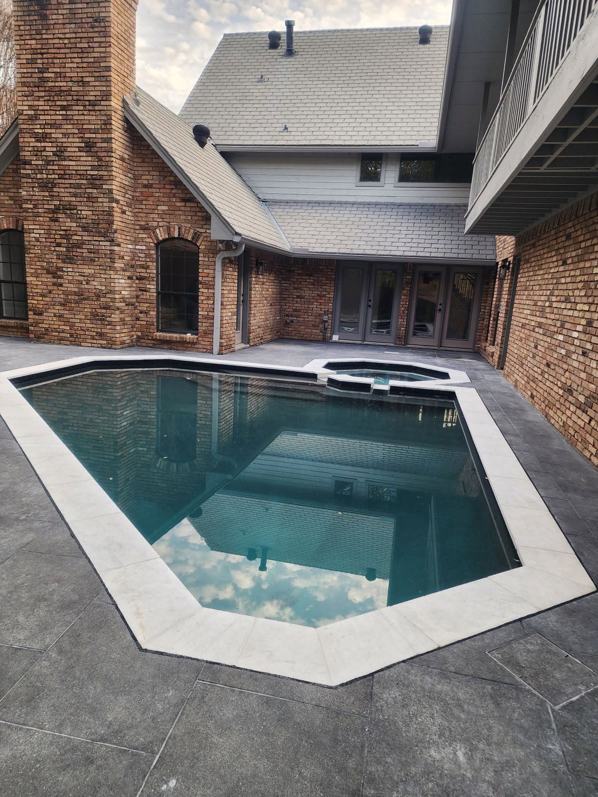 A recently remodeled residential pool featuring a modern octagonal shape with a smooth stone coping, surrounded by a textured dark paver deck. The pool's water reflects the sky and adjoining brick and wooden structures.