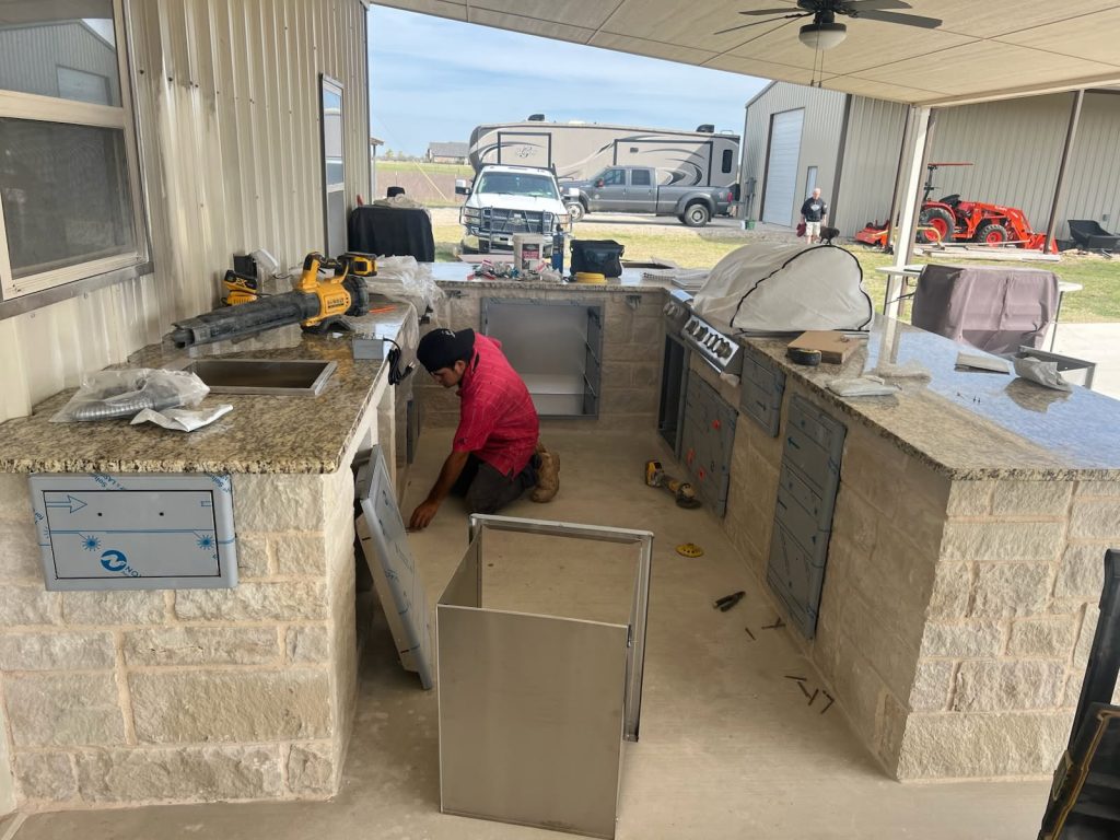 A worker assembling a custom outdoor kitchen with granite countertops and stainless steel appliances, set in a spacious outdoor area.