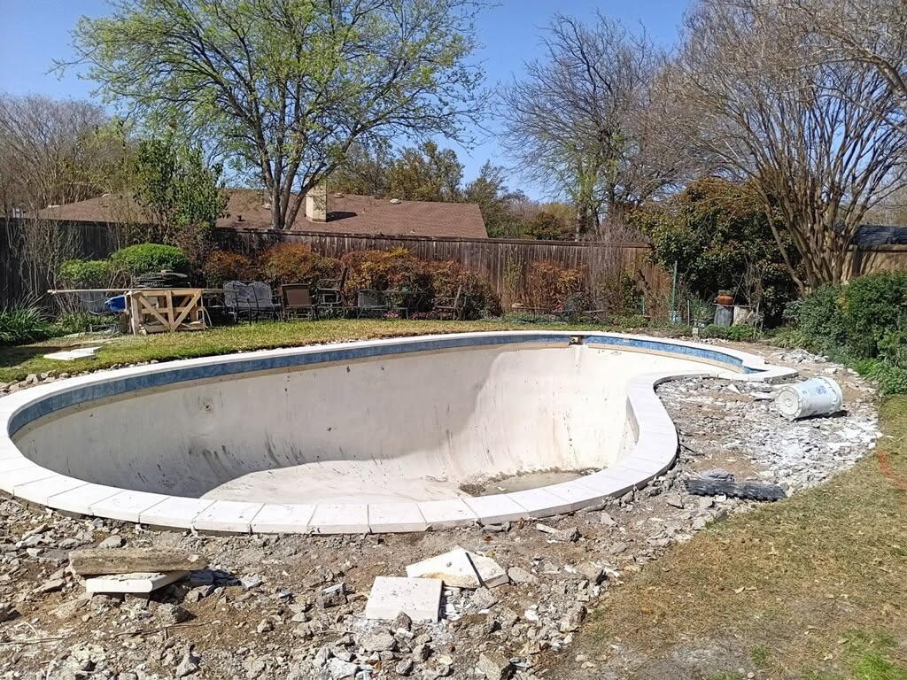 Before image of a residential pool undergoing renovations, showing an empty pool shell surrounded by debris and landscaping in a backyard setting.