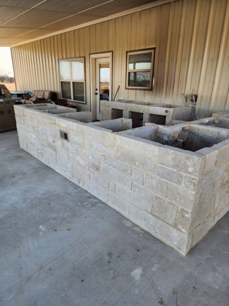 An unfinished outdoor kitchen structure made of stone blocks, with a metal building in the background and windows visible.