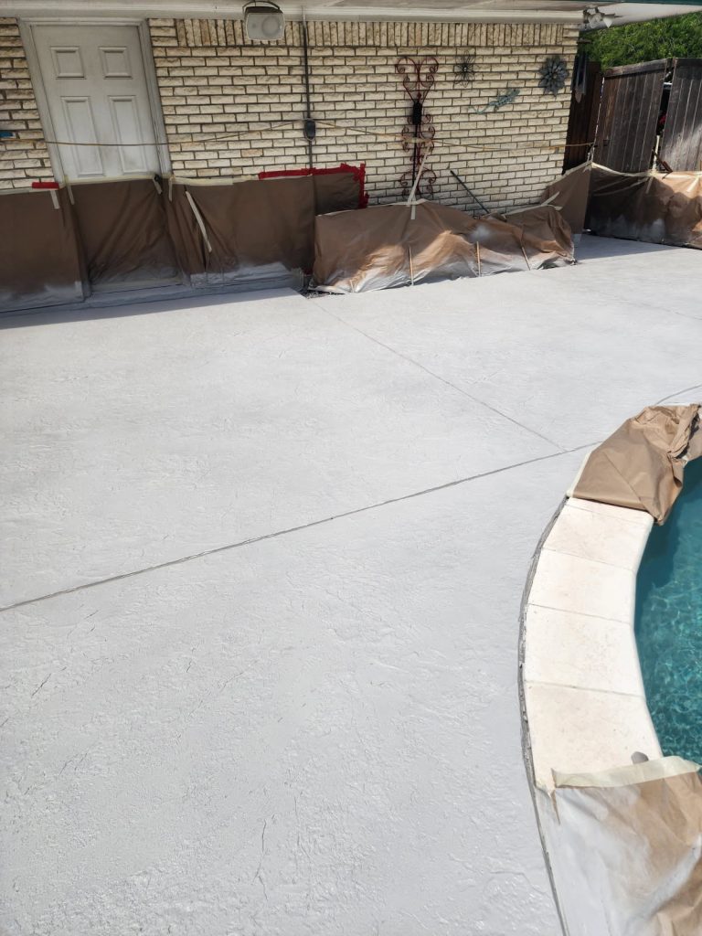 A residential pool area with a freshly resurfaced deck, featuring a smooth, light gray finish. The pool is partially visible, with a beige coping along the edge, while protective coverings are placed around the perimeter of the work area.