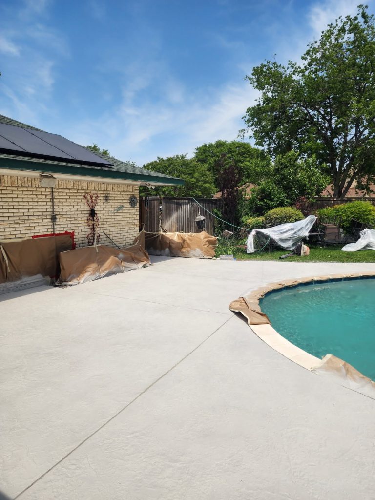 View of a backyard pool area undergoing remodeling, featuring a partially visible pool, freshly poured concrete deck, and outdoor landscaping with green trees and foliage.