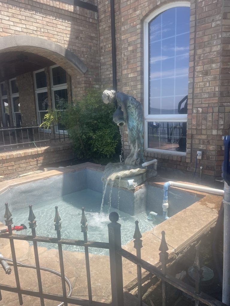 A close-up view of a pool area featuring a decorative fountain statue pouring water into a small pool, surrounded by brick walls and foliage.
