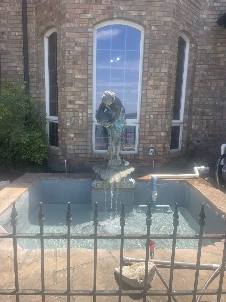 A decorative fountain featuring a statue of a woman pouring water into a small pool, surrounded by a wrought iron fence and brick walls.