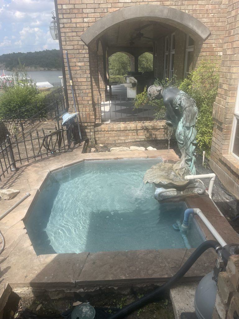 Small backyard pool with a statue fountain and clear blue water, surrounded by stone and brick architecture.