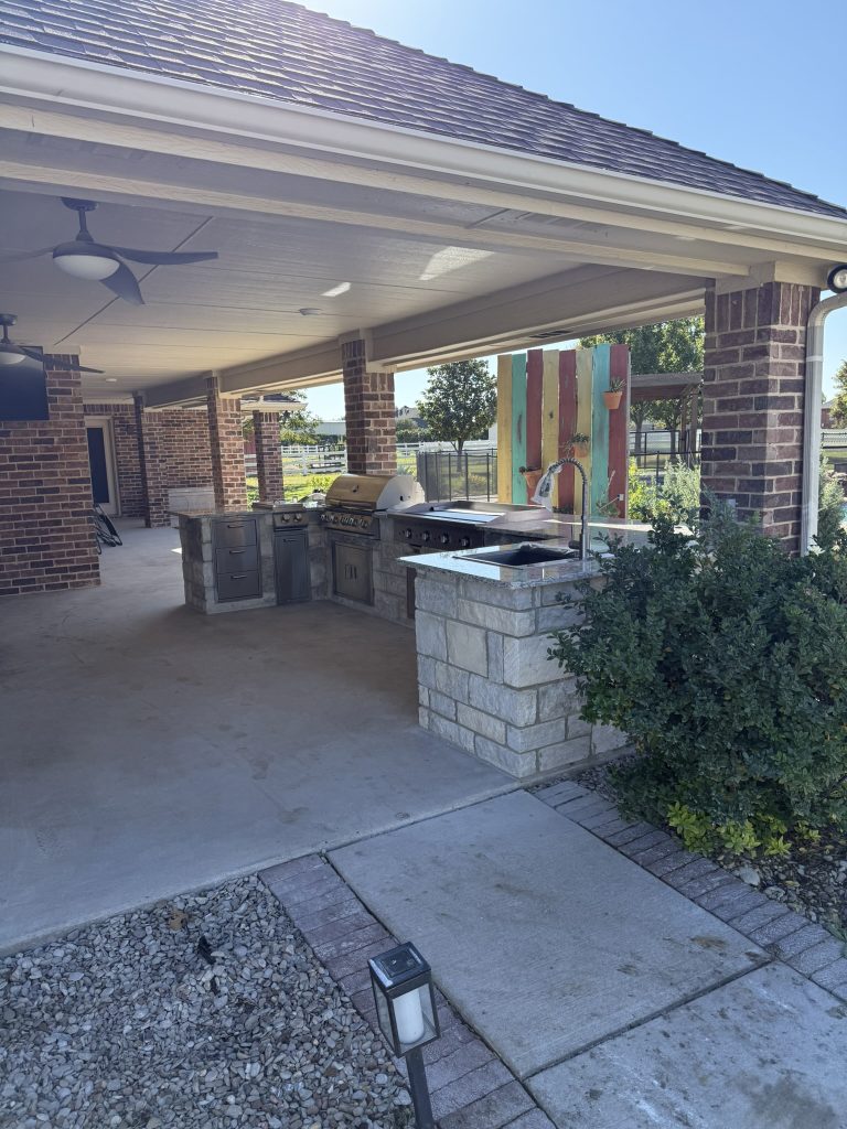 Outdoor kitchen area featuring a stone grill, countertops, and pendant lighting, set beneath a covered patio.
